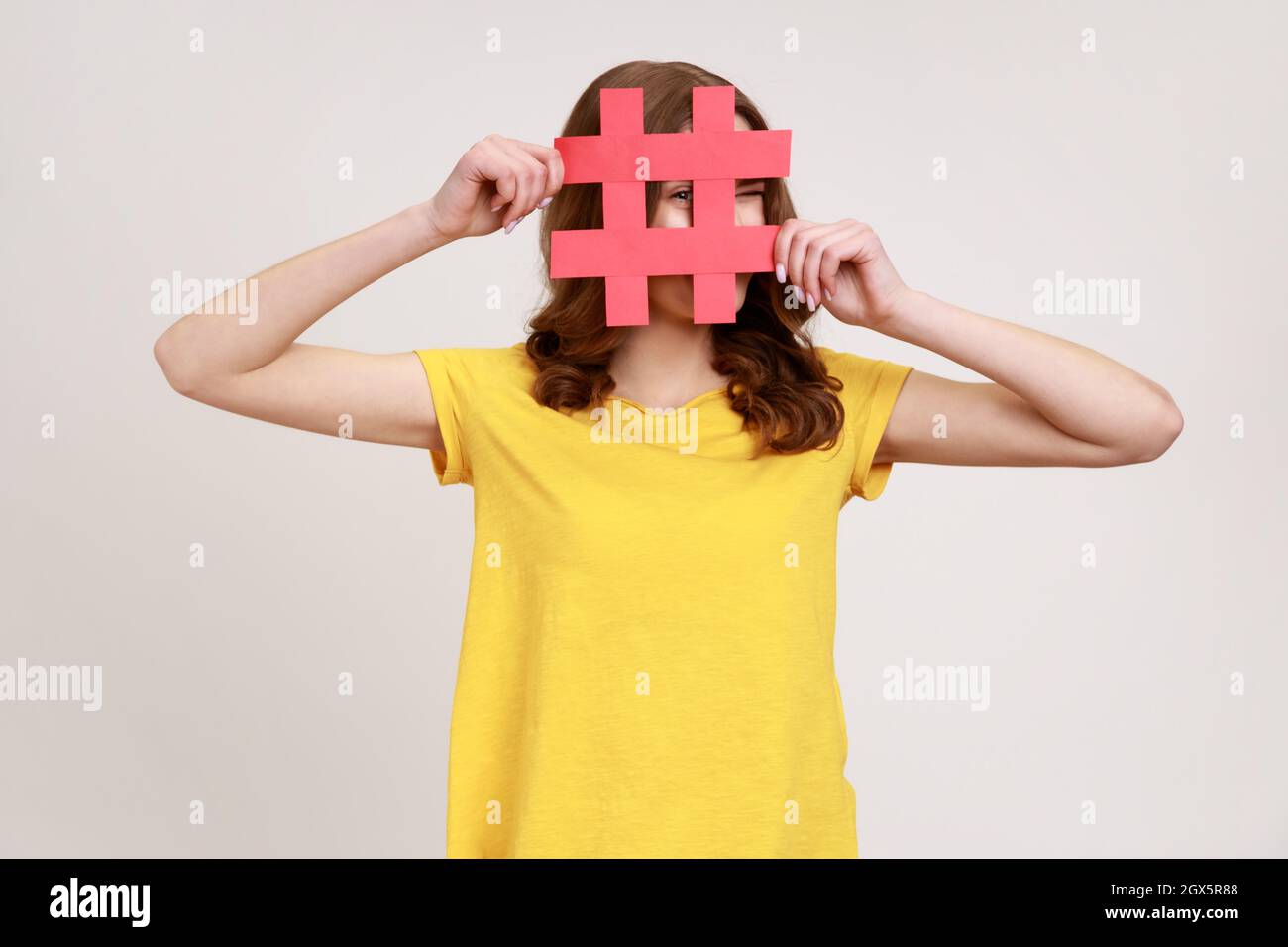 Neugierige, rothaarige Frau im jungen Alter in gelbem T-Shirt, die durch rotes Hashtag-Schild schaut, nach richtigen Beiträgen in sozialen Medien sucht, ausspioniert. Innenaufnahme des Studios isoliert auf grauem Hintergrund. Stockfoto