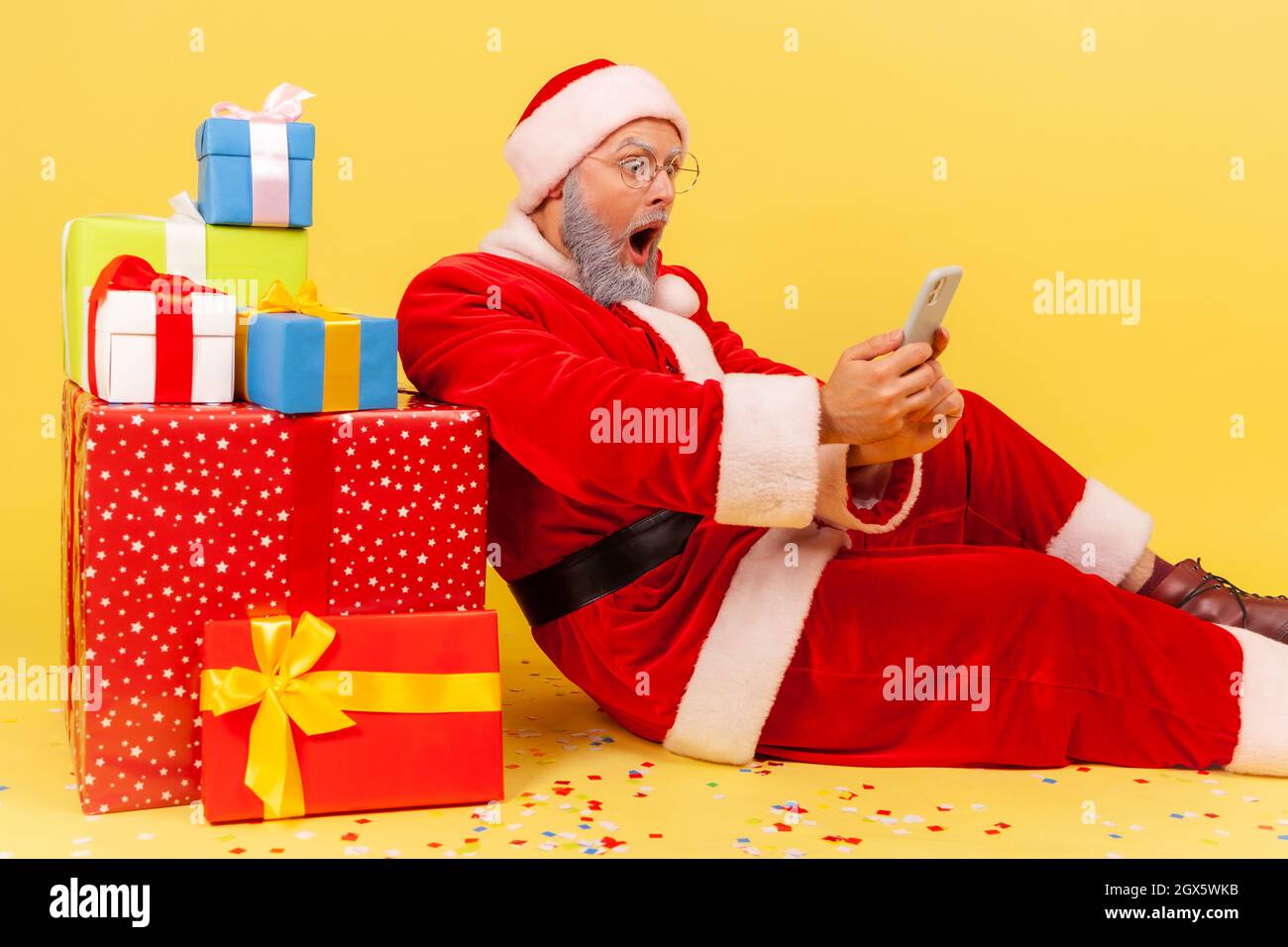 Seitenansicht eines älteren Mannes mit grauem Bart, der das weihnachtsmann-Kostüm trägt, der in der Nähe von Neujahrsgeschenken sitzt und mit dem Smartphone eine schockierende Nachricht liest. Innenaufnahme des Studios isoliert auf gelbem Hintergrund. Stockfoto