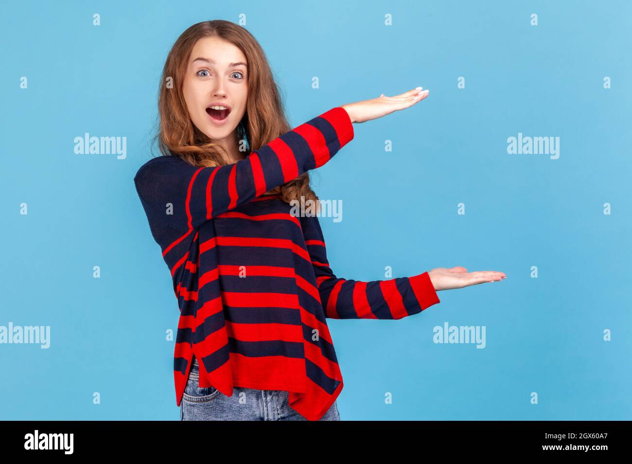 Extrem erstaunt Frau trägt gestreiften lässigen Stil Pullover, präsentiert Bereich zwischen den Händen für Werbung, zeigt riesige Größe. Innenaufnahme des Studios isoliert auf blauem Hintergrund. Stockfoto