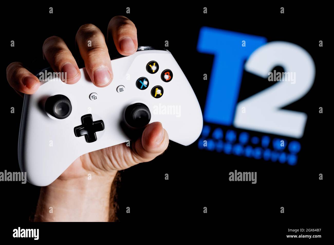 Ein weißes Gamepad, das in der Hand auf dem schwarzen Hintergrund mit dem Logo von Take-Two gehalten ist. Stockfoto