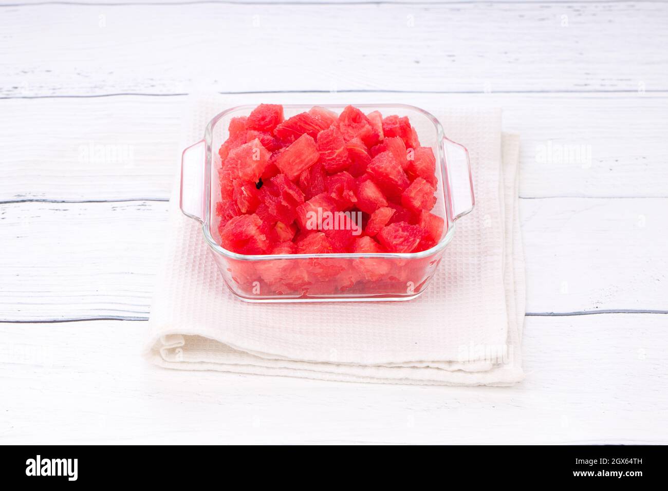 Wassermelonenscheiben, Ausschnitt aus Wassermelone, in einer transparenten quadratischen Glasschale mit weißem rustikalem Hintergrund angeordnet, isoliert. Stockfoto