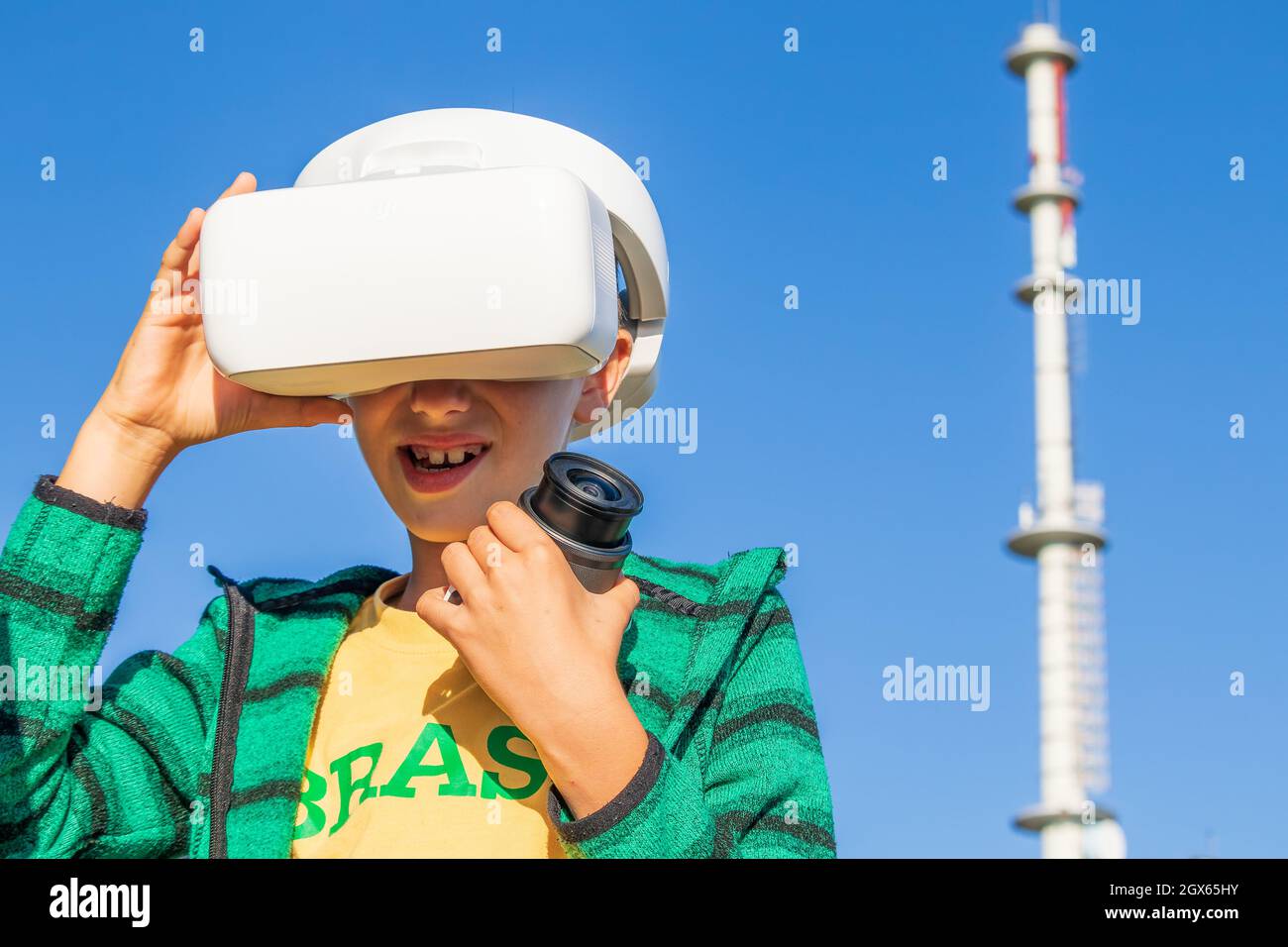 Kind trägt eine Virtual-Reality-Brille und hält die Kamera in der linken Hand, im Hintergrund ist der blaue klare Himmel und das Mobilfunkrelais zu sehen Stockfoto