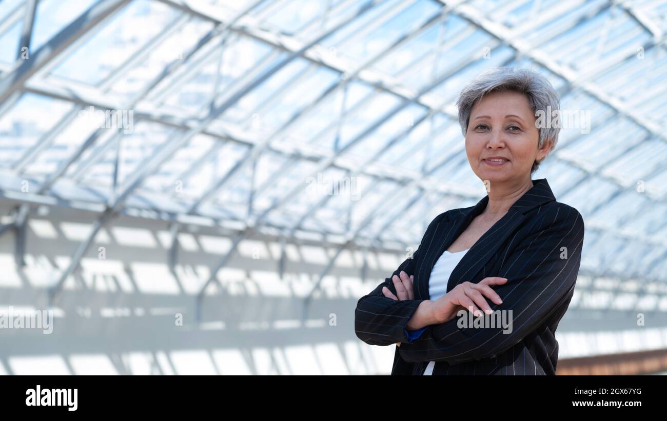 Glückliche erfolgreiche reife asiatische Geschäftsfrau posiert mit Armen im Geschäftsgebäude gefaltet Stockfoto