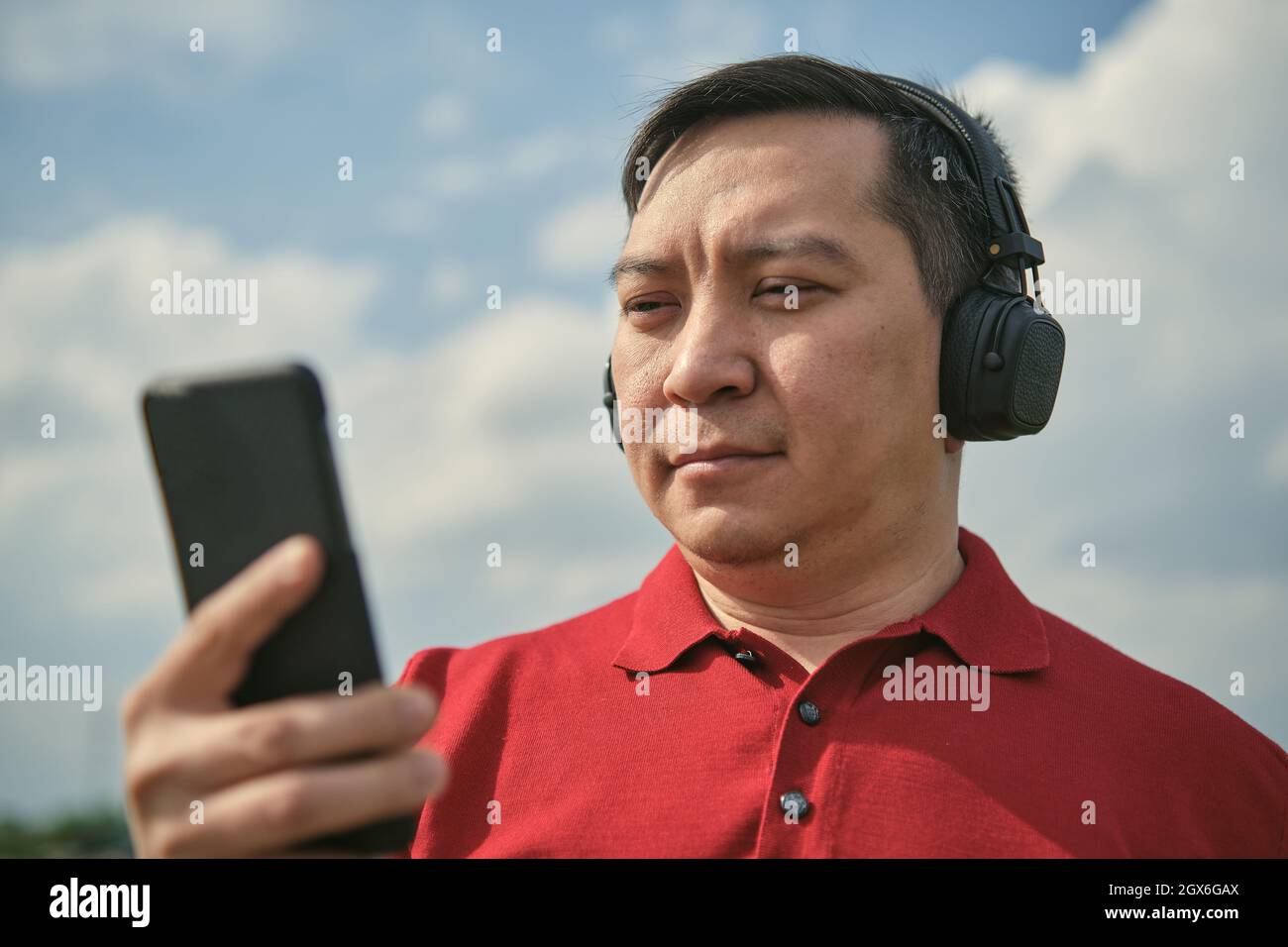 Ein asiatischer Mann mittleren Alters mit Kopfhörern im Freien, der vor dem Hintergrund des Himmels Musik hört, Mobiltelefon in der Hand Stockfoto