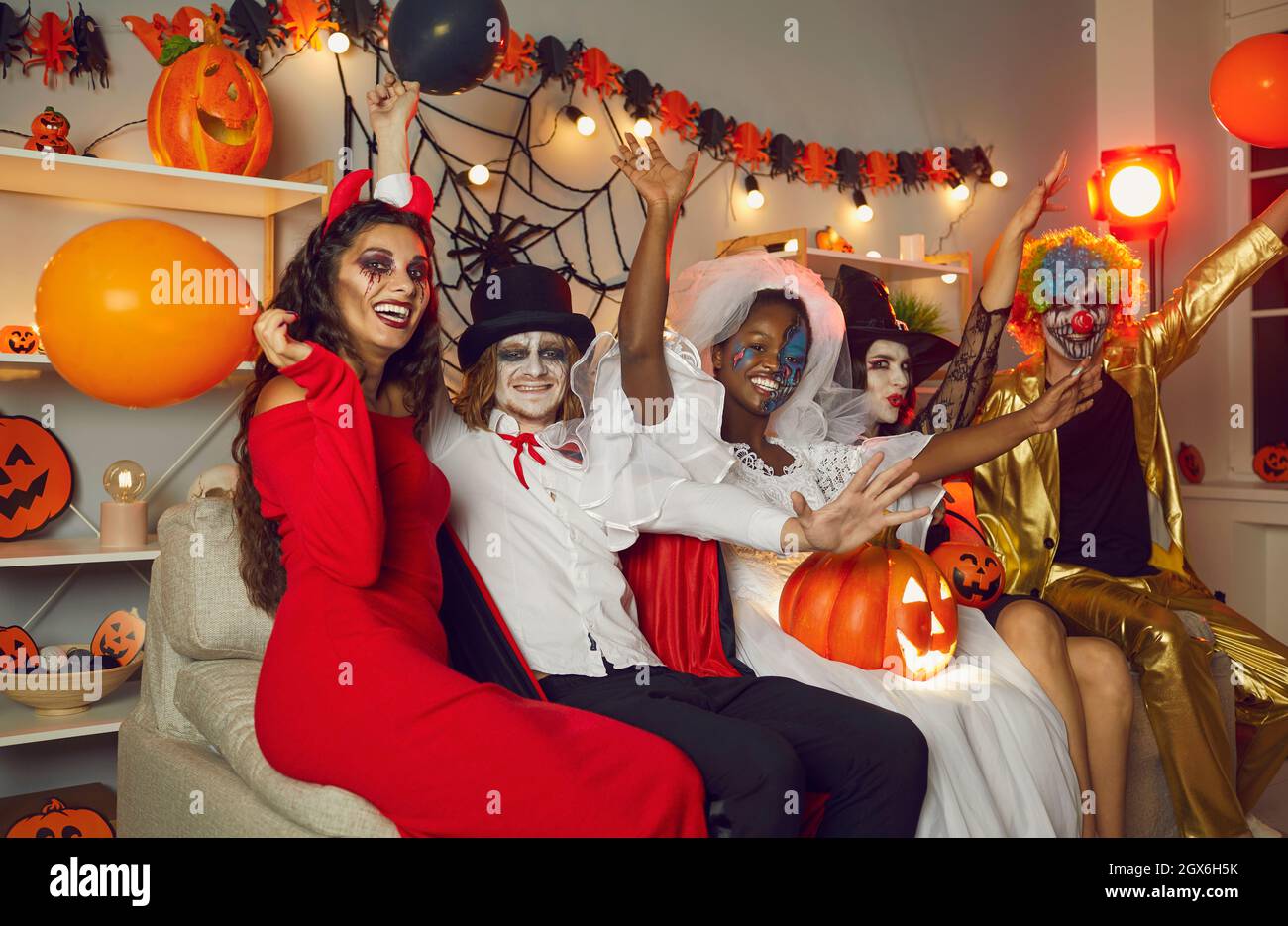 Fröhliche Freunde in gruseligen Kostümen sitzen alle zusammen auf der Couch auf der Halloween Party Stockfoto