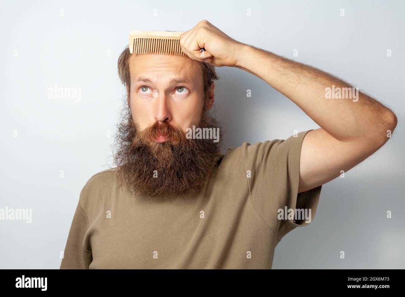 Porträt eines erwachsenen kahlen Mannes mit Haarbürste, Alopezie leidend, mit verwirrtem Gesichtsausdruck aufblickend, bärtiger Mann mit T-Shirt. Innenaufnahme des Studios isoliert auf grauem Hintergrund. Stockfoto