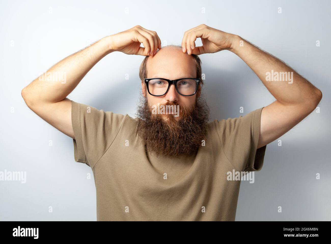 Mann berührt seinen Kopf. Problem mit Haaren, leiden Alopezie, braucht Trichologe Beratung, kahler bärtiger Mann trägt T-Shirt und Brille. Innenaufnahme des Studios isoliert auf grauem Hintergrund. Stockfoto