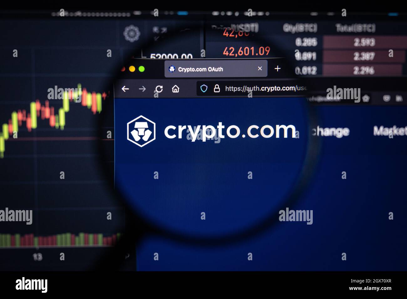 Crypto.com Firmenlogo auf einer Website mit verschwommenen Börsenentwicklungen im Hintergrund, auf einem Computerbildschirm durch eine Lupe gesehen Stockfoto