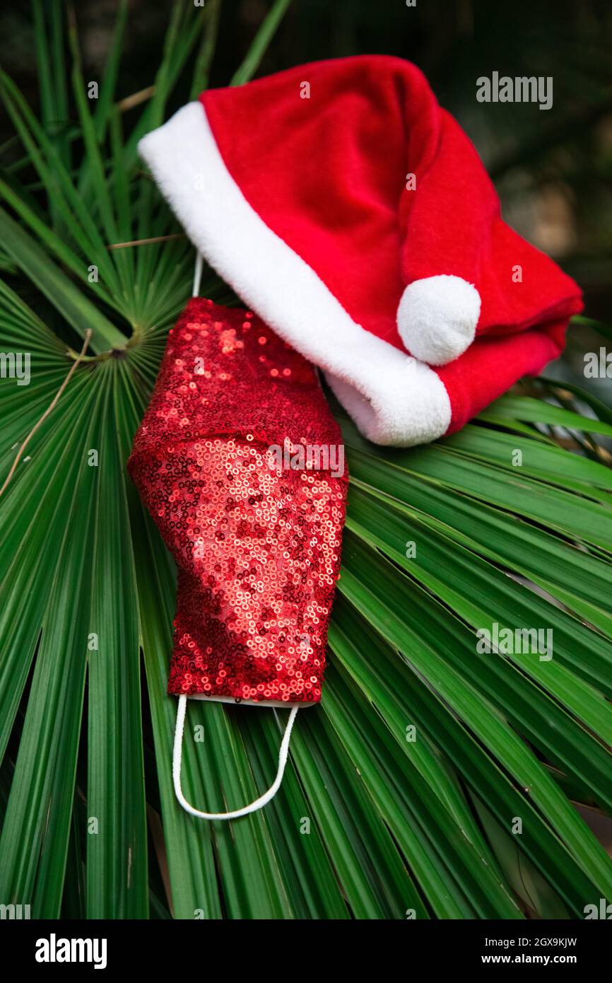Roter Weihnachtshut mit weißem Rand, rote schützende medizinische funkelnde Maske auf grünem Palmblatt. Zubehör für die Neujahr-Party 2021. Wiederverwendbare covid Stockfoto