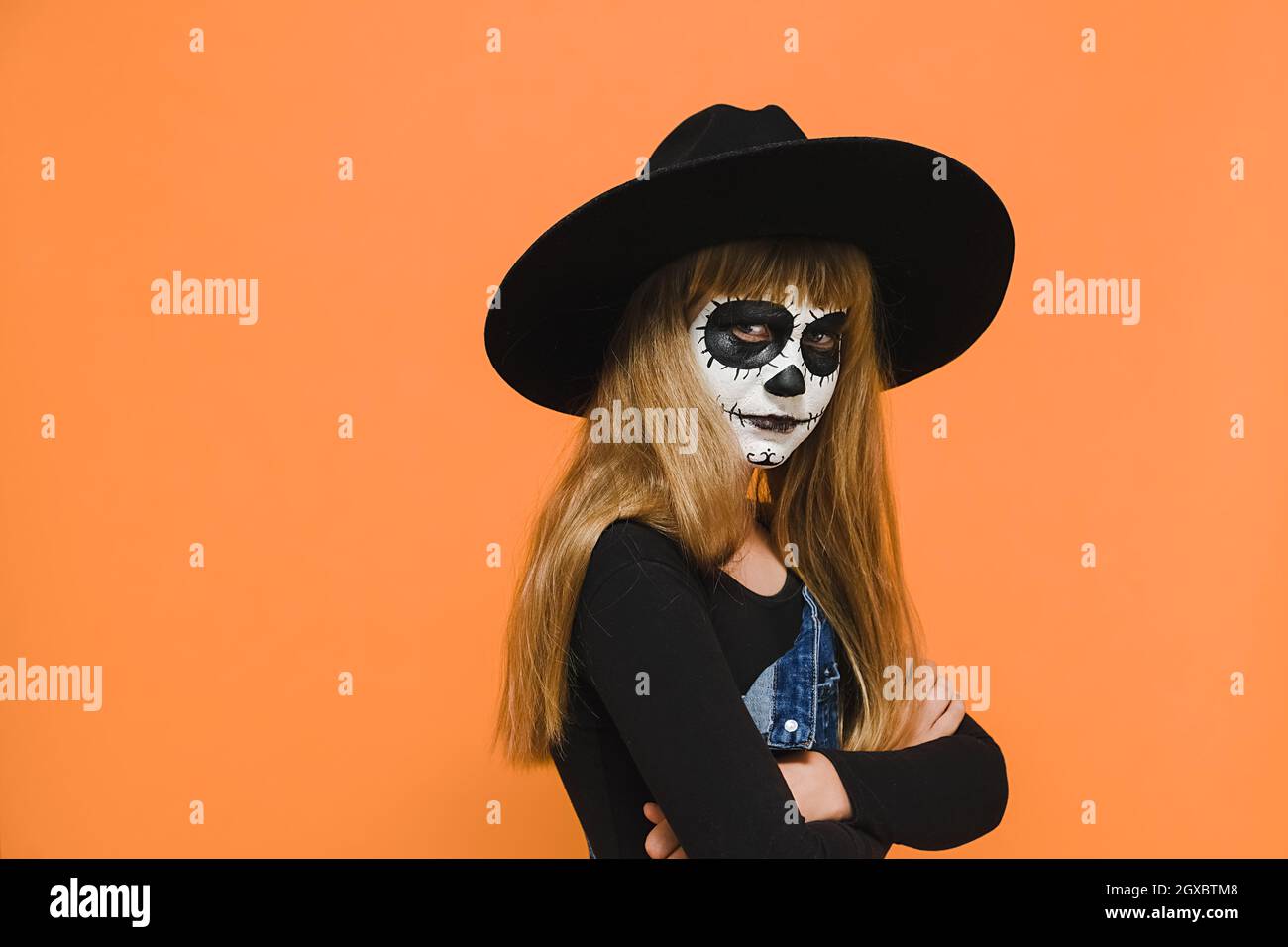 Wütend traurig kleine blonde Mädchen Kind mit Halloween gruselige Make-up-Maske, trägt schwarzen Hut, gekreuzte Arme, unglücklich Blick auf die Kamera Stockfoto