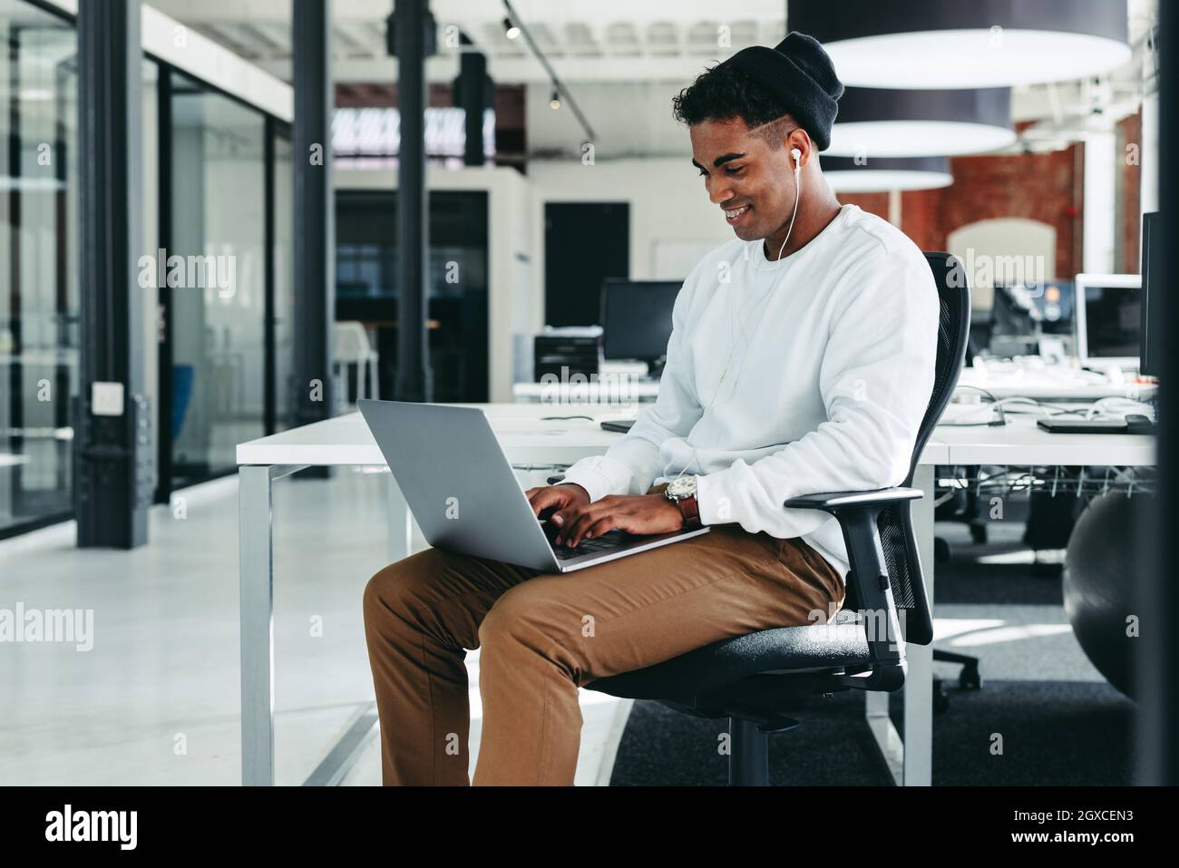 Glücklicher junger Geschäftsmann, der im Büro an einem neuen Projekt arbeitet. Kreativer Geschäftsmann, der fröhlich lächelt, während er an einem modernen Arbeitsplatz einen Laptop benutzt. Ja Stockfoto