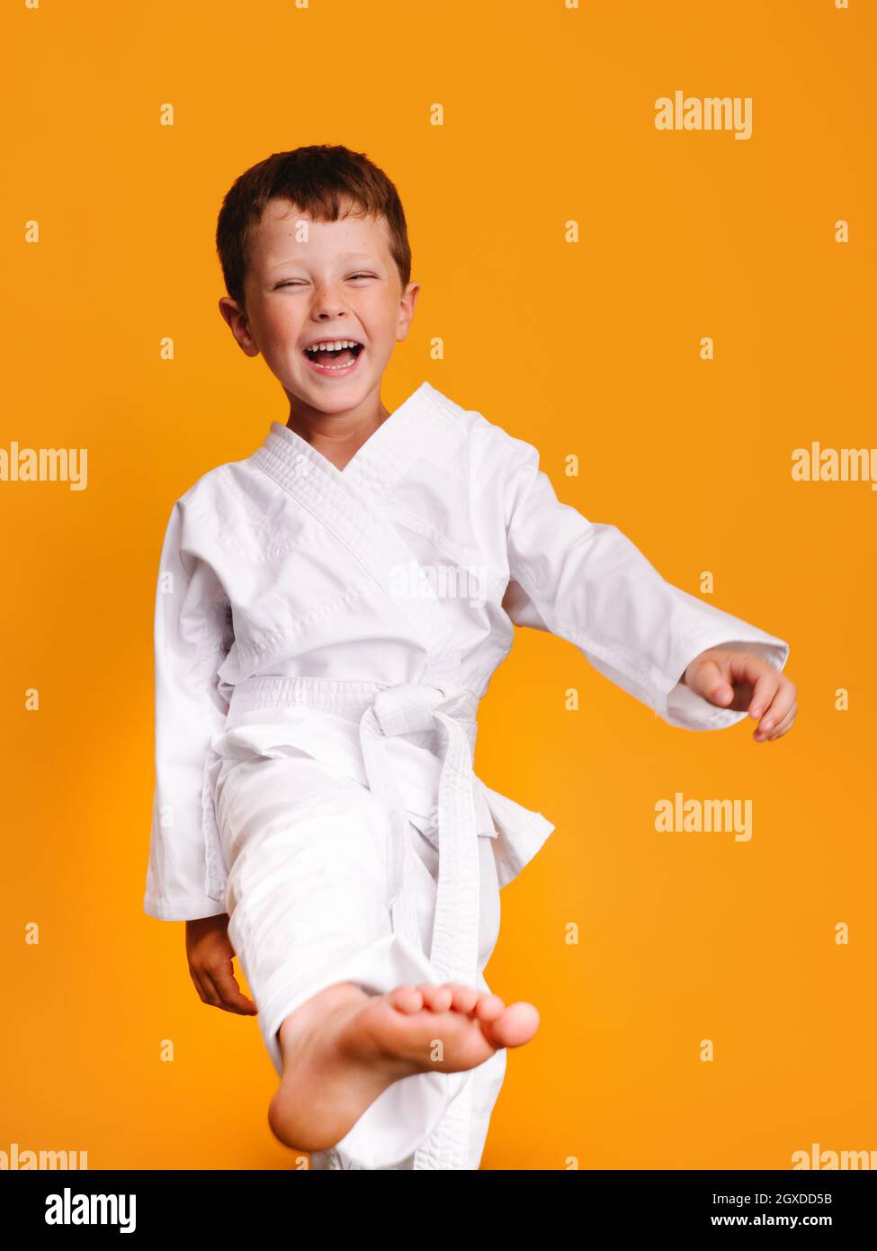 Vorderansicht eines lächelnden Kindes in einem weißen Kimono mit halb geschlossenen Augen, das rechte Bein und den rechten Fuß in einer Studiosession mit orangefarbenem Rücken hebt Stockfoto