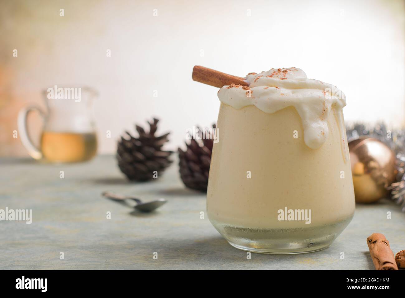 Glas Milchpunsch mit Zimtpulver auf geschlagenen Eiweiß gegen Pinienzapfen am Weihnachtstag auf hellem Hintergrund Stockfoto