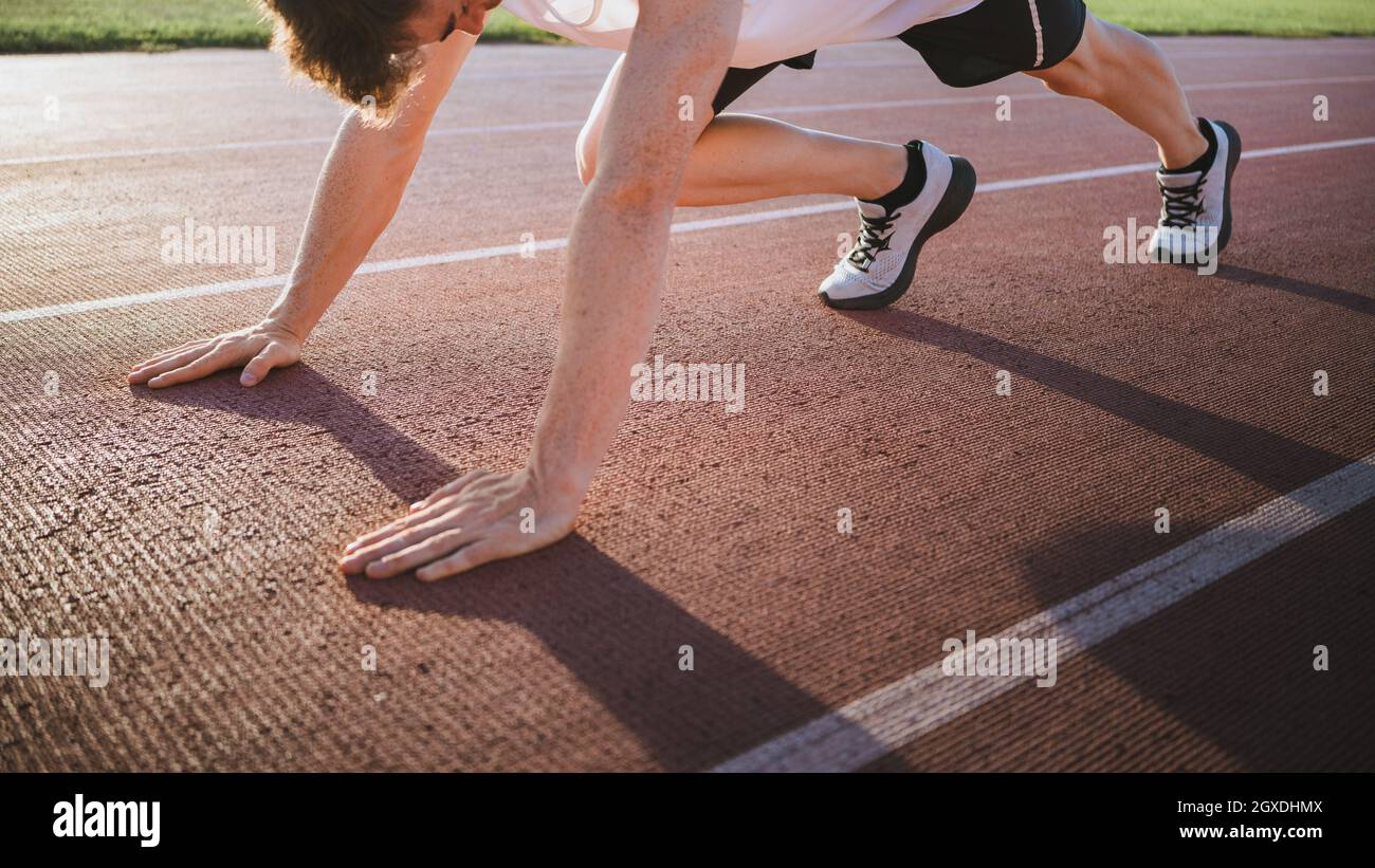 Crop männlichen Athleten in Turnschuhen in Startposition stehen vor dem Training auf der Strecke in Sonnenlicht Stockfoto