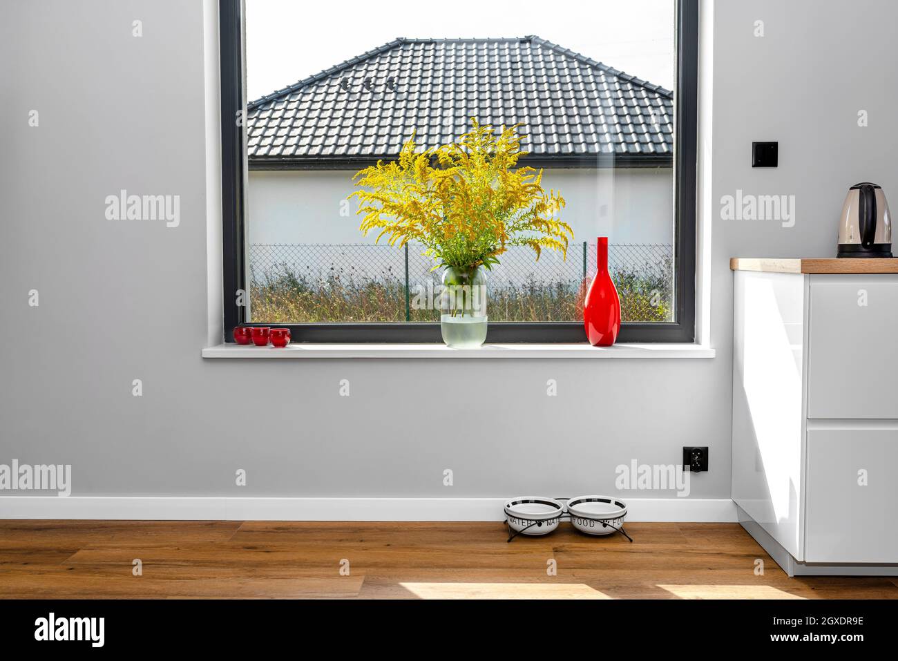 Ein großes Fenster im Esszimmer, das mit der Küche verbunden ist, auf der Fensterbank befindet sich eine rote Vase mit Goldrute, sichtbare Hundeschüsseln und ein Schrank. Stockfoto