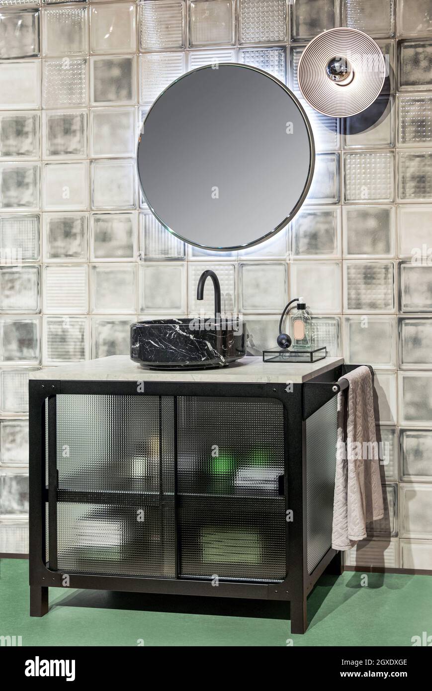 Moderne Einrichtung des Badezimmers mit Waschbecken aus schwarzem Marmor und beleuchtetem runden Spiegel, der an der gefliesten Wand hängt Stockfoto