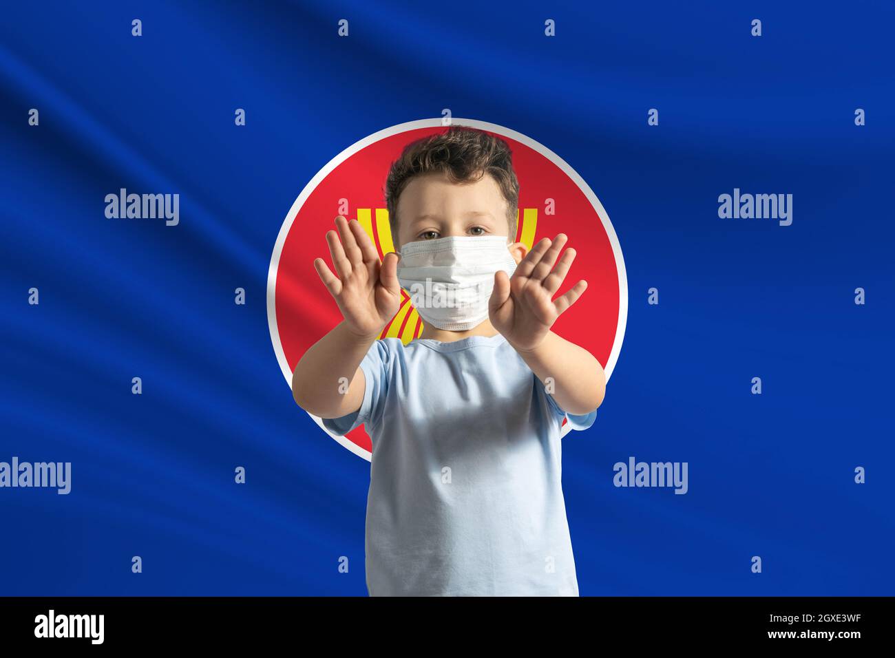 Kleiner weißer Junge in einer Schutzmaske auf dem Hintergrund der Flagge der Vereinigung südostasiatischer Nationen. Macht mit seinen Händen ein Stoppschild, bleiben Stockfoto