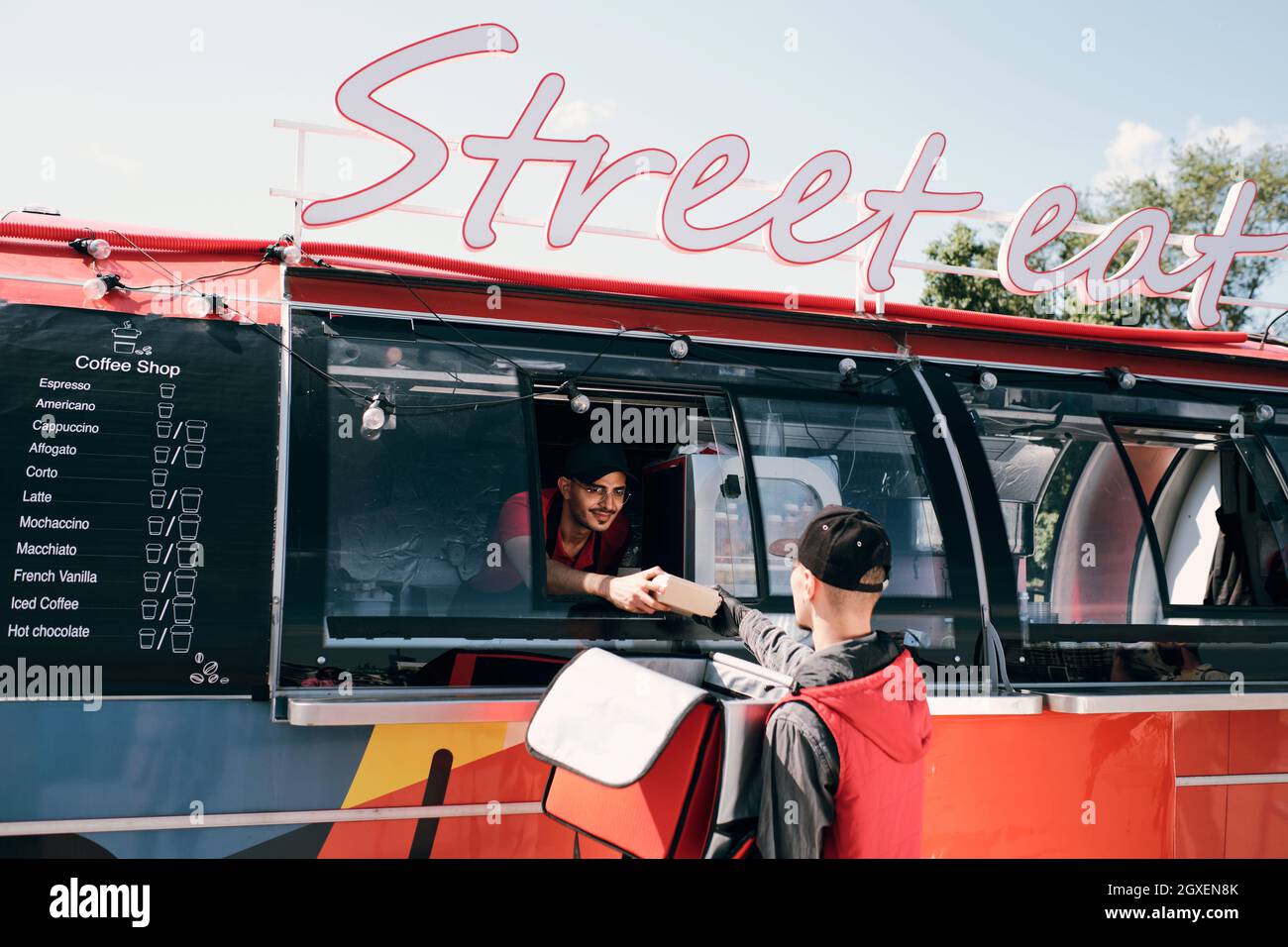 Junger männlicher Angestellter des Straßenlebensmittelfahrzeugs, der verpackte Schachtel mit Snack an einen Kurier mit großer Tasche übergab Stockfoto