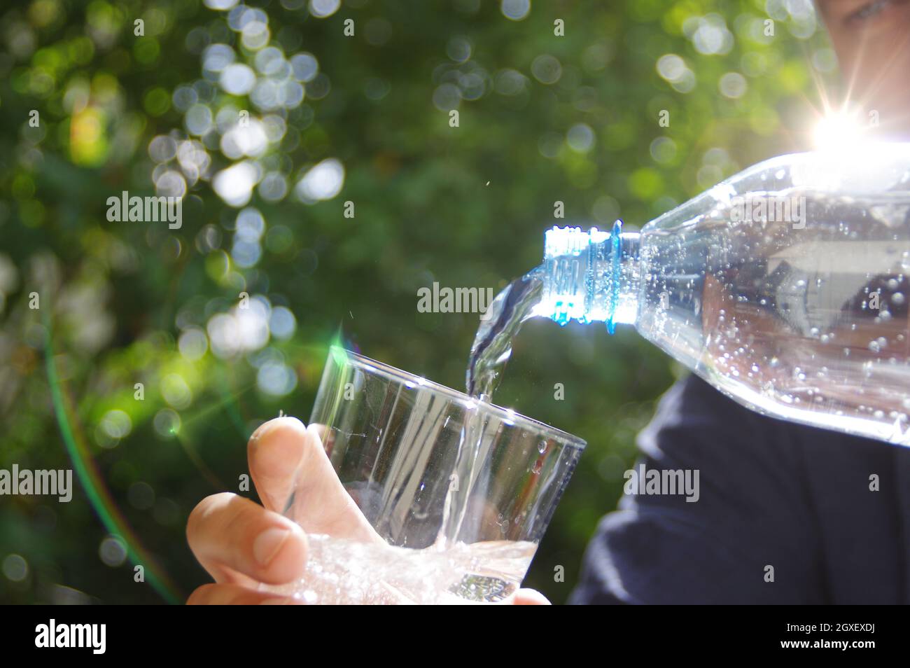 Wasser in ein Glas in der Hand mit Garten im Hintergrund gegossen. Erfrischung mit sauberem, natürlichem Mineralwasser aus einer Plastikflasche. Stockfoto