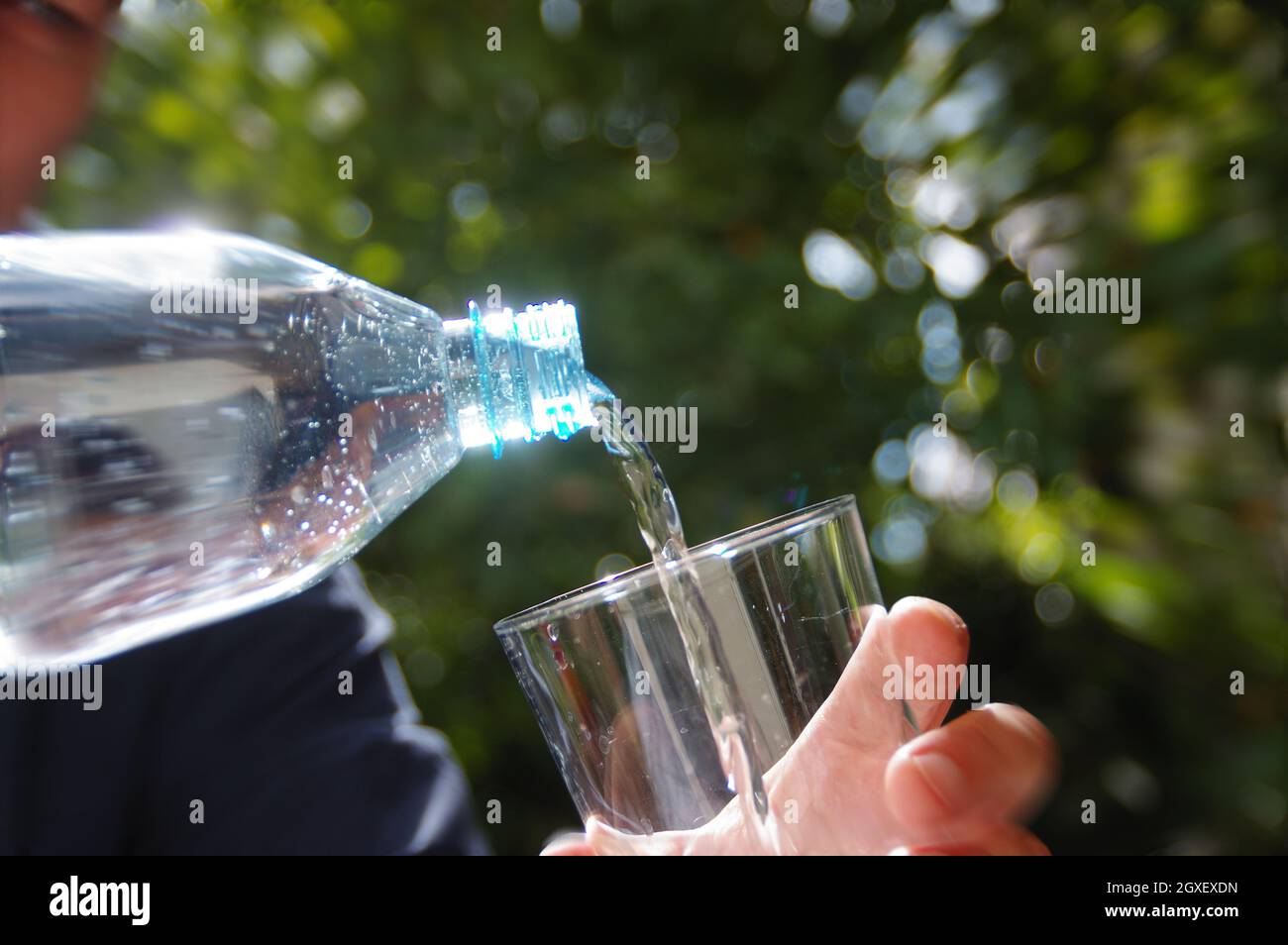Wasser in ein Glas in der Hand mit Garten im Hintergrund gegossen. Erfrischung mit sauberem, natürlichem Mineralwasser aus einer Plastikflasche. Stockfoto
