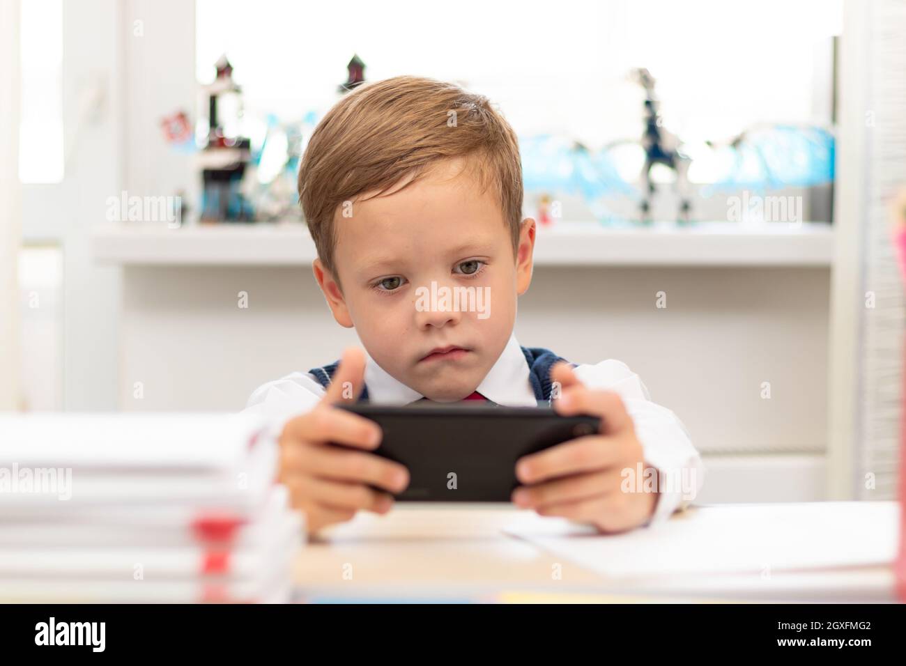 Ein süßer Erstklässler-Junge in Schuluniform auf hellem Hintergrund sitzt zu Hause mit einem Smartphone in der Hand an einem Schreibtisch. Selektiver Fokus. Nahaufnahme. Po Stockfoto