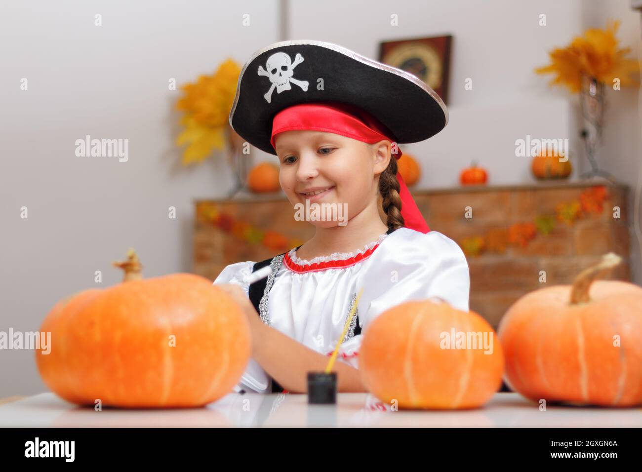 Halloween zu Hause feiern. Ein kleines Mädchen, das als Pirat gekleidet ist, sitzt am Tisch. Das Kind bereitet sich auf die Party vor und zieht den Kürbis an. Stockfoto