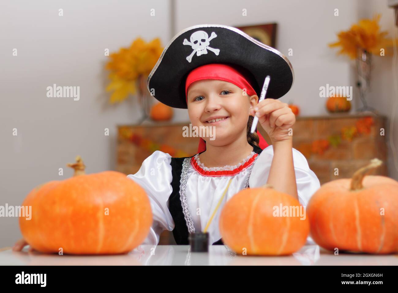 Halloween zu Hause feiern. Ein kleines Mädchen, das als Pirat gekleidet ist, sitzt am Tisch und zieht einen Kürbis an. Das Kind schaut in die Kamera. Stockfoto