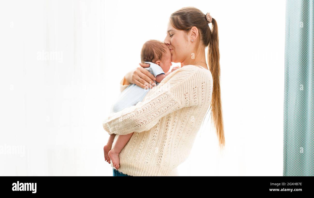 Glücklich lächelnde junge Mutter kuscheln und küssen ihren neugeborenen Jungen gegen helle Sonne scheint durch Fenster. Konzept von Familie Glück und Liebe Stockfoto