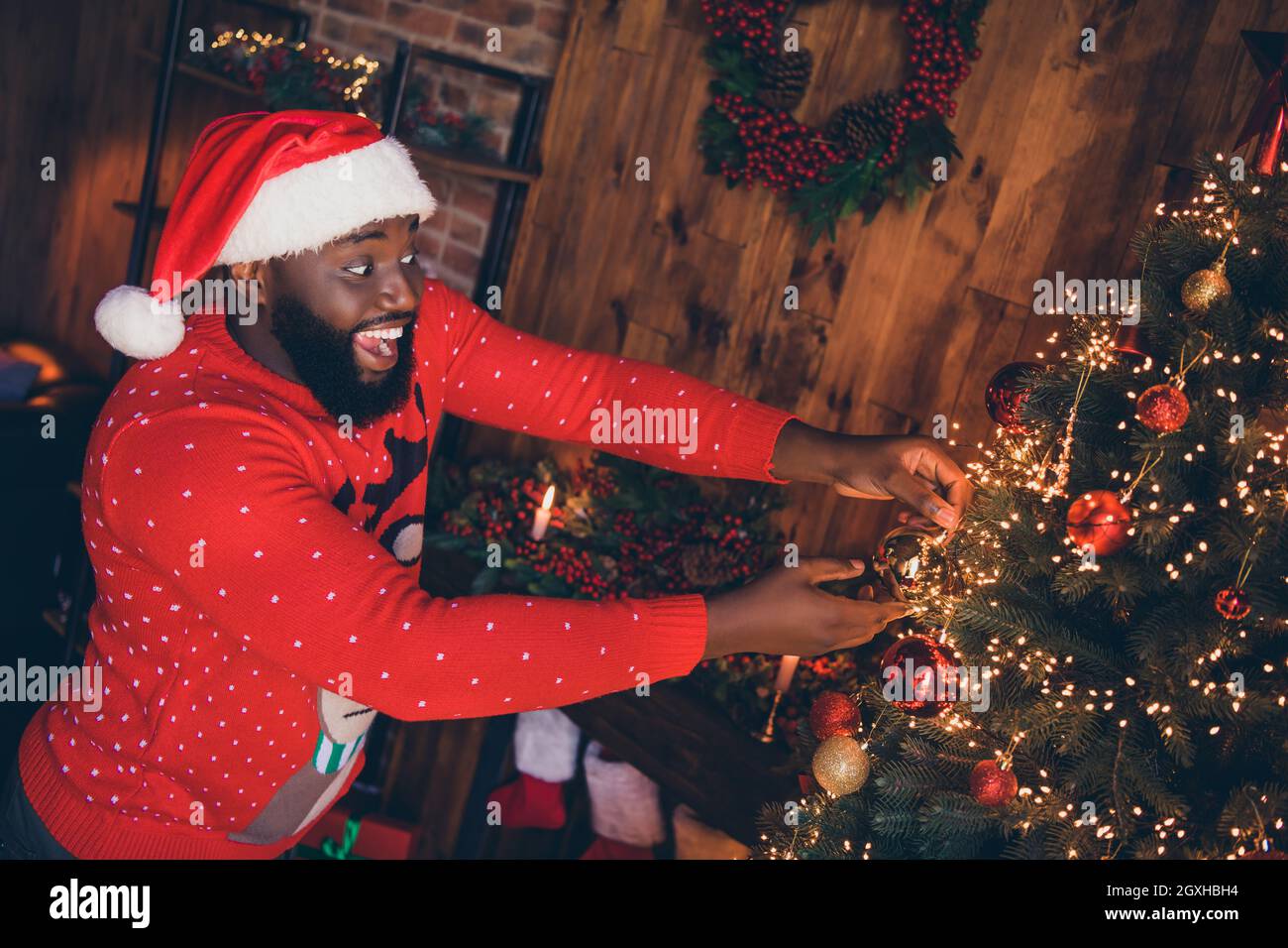 Profilfoto von funky junge Brunet Kerl setzen Dekoration tragen rote Mütze Pullover im Loft zu Hause neues Jahr Stockfoto
