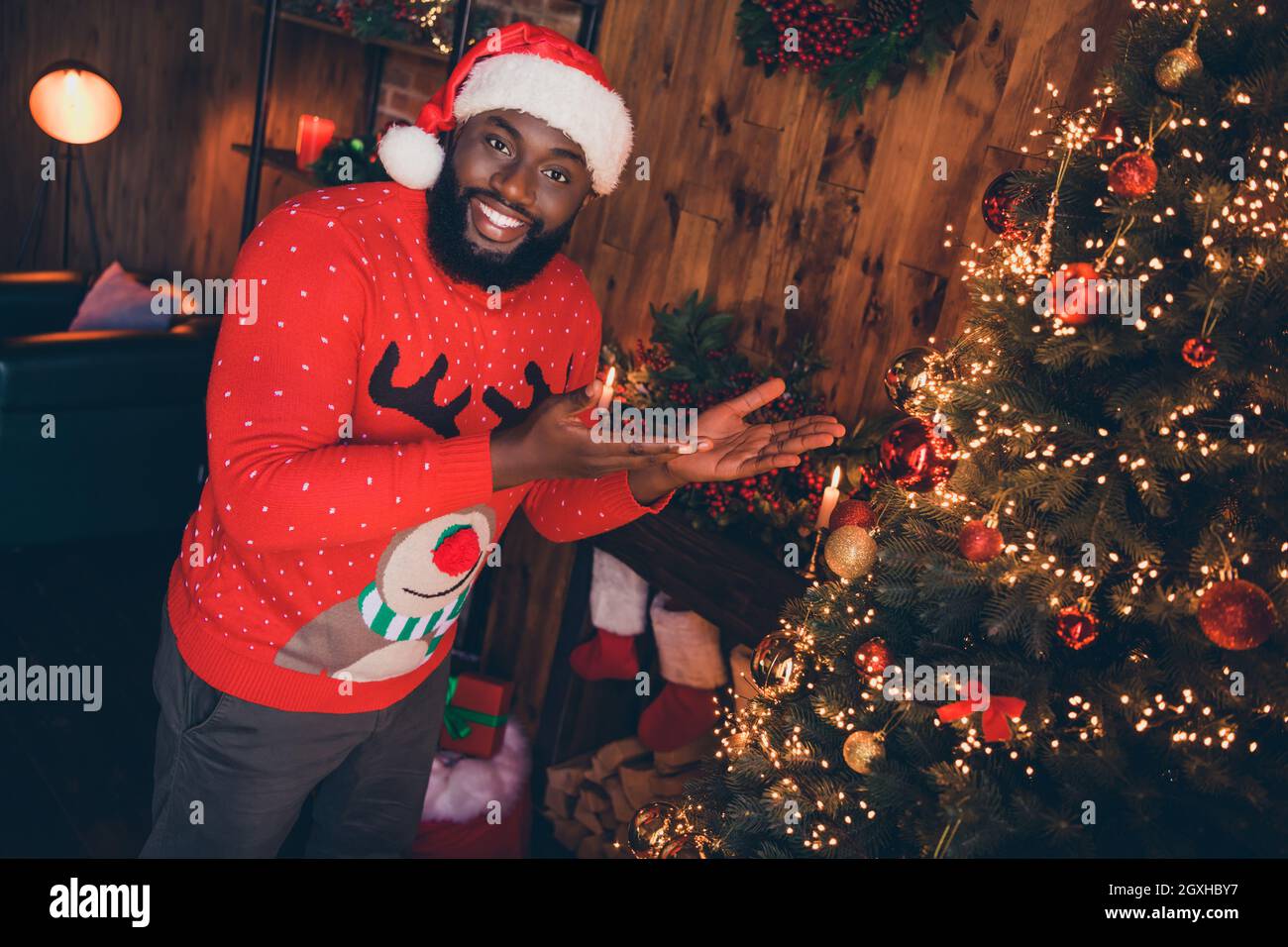 Foto von schönen Millennial Brunet Kerl Point Tree tragen rote Mütze Pullover im Loft zu Hause Neujahr Stockfoto