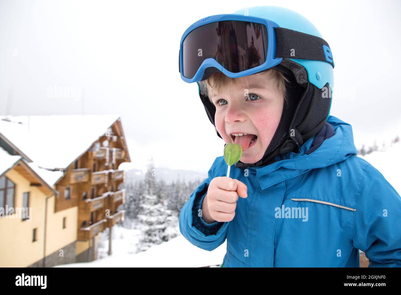 Niedlicher Junge im Helm, Brille und blauen Overall im Skigebiet. Essen Süßigkeiten auf einem Stock, eine Minute Ruhe. Aktive Winterunterhaltung für Chil Stockfoto