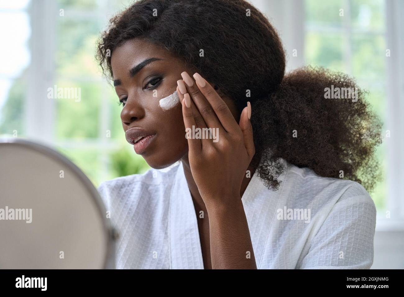 Attraktive schwarze junge Frau, die Creme auf die Gesichtshaut auftragen, die im Spiegel aussieht. Stockfoto