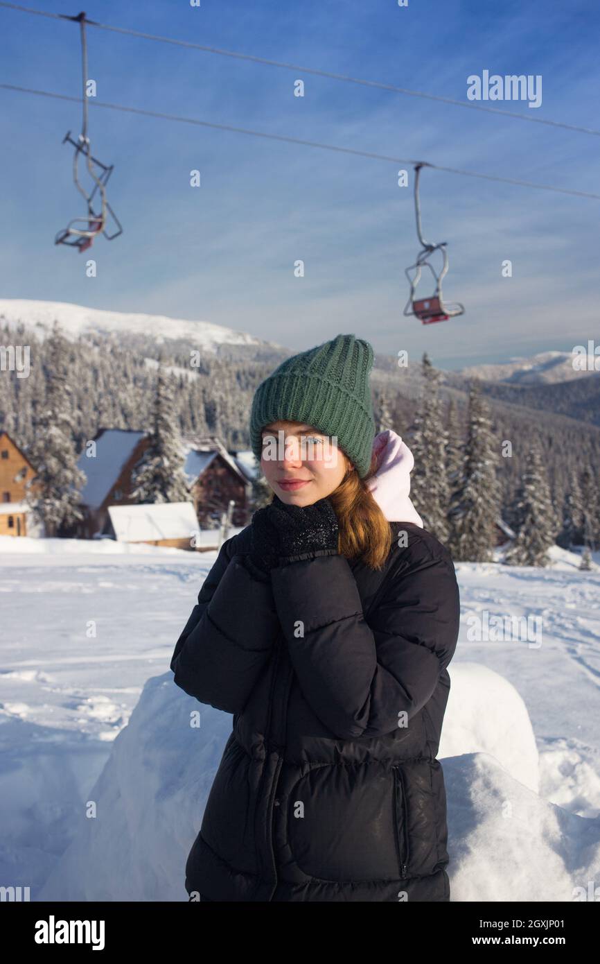 Nettes lächelndes Teenager-Mädchen in guter Laune steht vor dem Hintergrund von schneebedeckten Tannenbäumen und einem Skilift in einem Skigebiet. Kalter sonniger Tag. winte Stockfoto