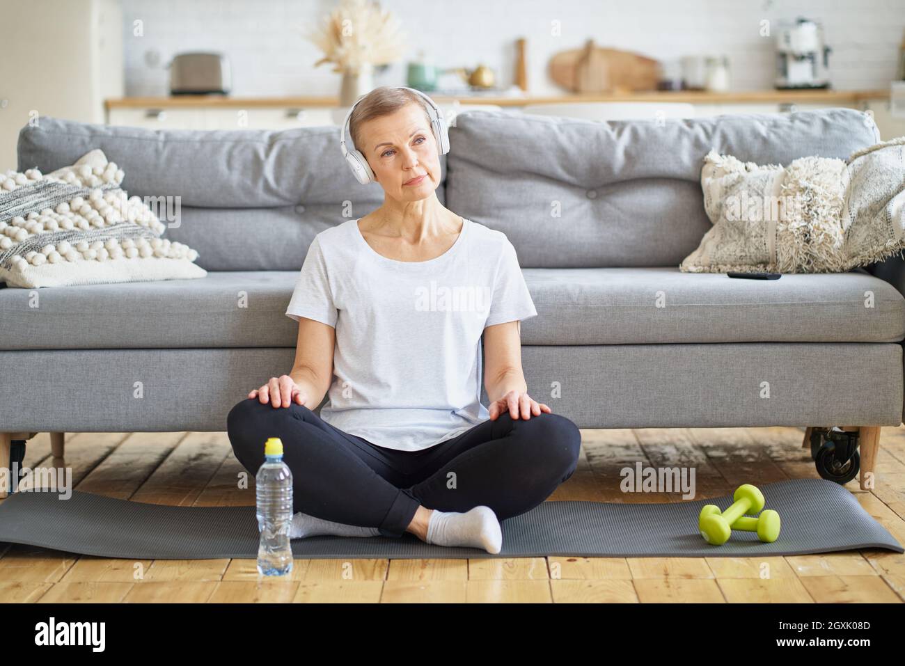 Ältere Frau sitzt mit gekreuzten Beinen in Kopfhörern und hört im Wohnzimmer Musik. Entspannen Sie sich nach dem Fitnesstraining. Stockfoto