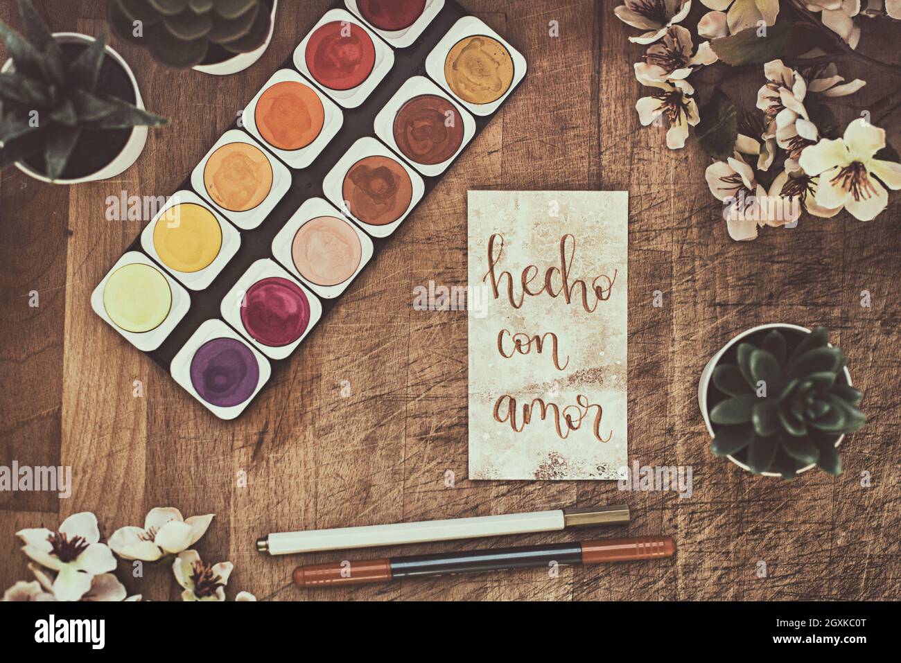 Aquarell malt auf einem Tisch mit Topfpflanzen, Blumen und einer Karte mit den Worten hecho con amor Stockfoto
