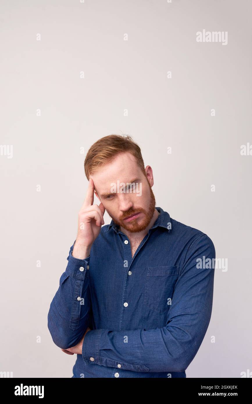 Ernsthafter junger Rotschopf-Mann mit Bart, der sich auf Gedanken konzentriert, die die Stirn vor grauem Hintergrund berühren Stockfoto