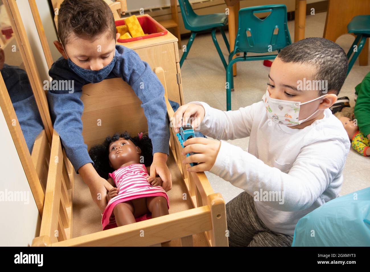 Bildung Vorschule 3-4-jährige zwei Jungen spielen im Familienbereich mit Puppe und Puppenbett, einer spielt mit Spielzeugeisenbahn, trägt Gesichtsmasken zum Schutz Covid Stockfoto