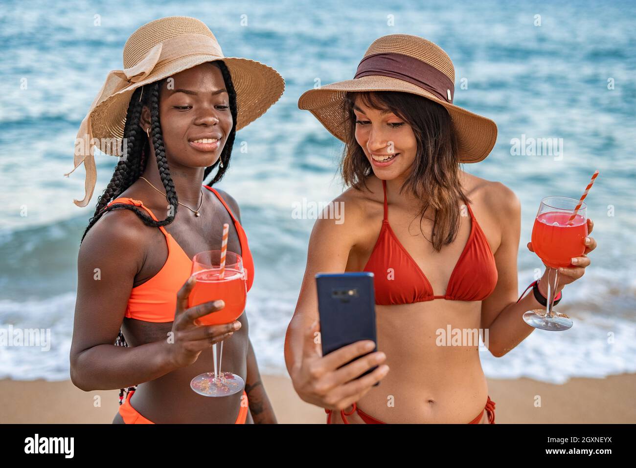 Stilvolle, fröhliche junge, vielfältige Freundinnen in Bikinis und Hüten, die Brille mit roten Getränken halten und Selfie auf dem Handy machen, während sie am Sommertag gemeinsam am Meer chillen Stockfoto