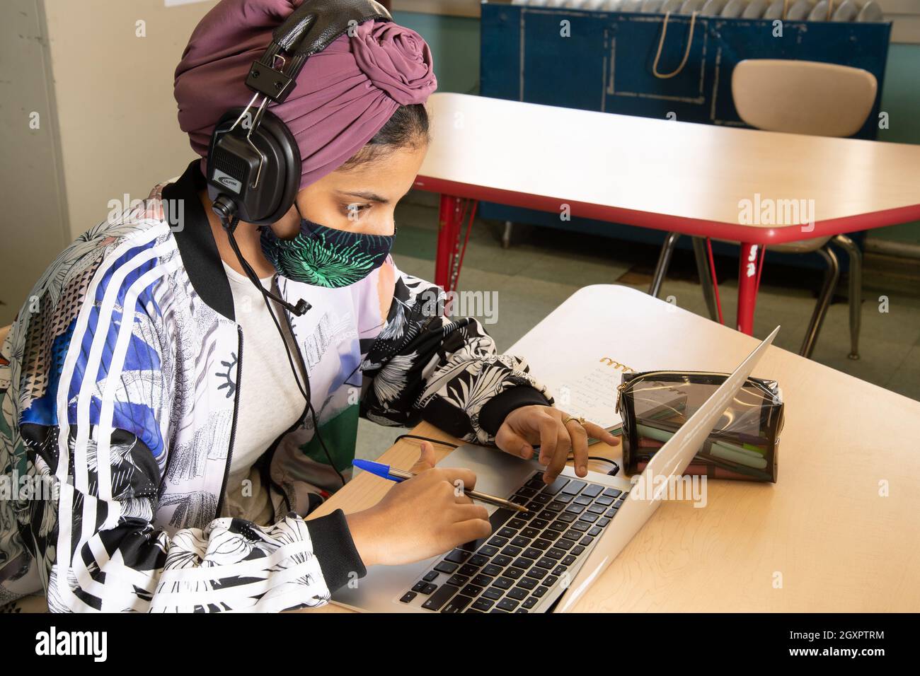 Schulschülerin, die in der Klasse an einem Laptop arbeitet, Kopfhörer trägt und eine Gesichtsmaske trägt, um sich vor dem Covid-19-Virus zu schützen Stockfoto