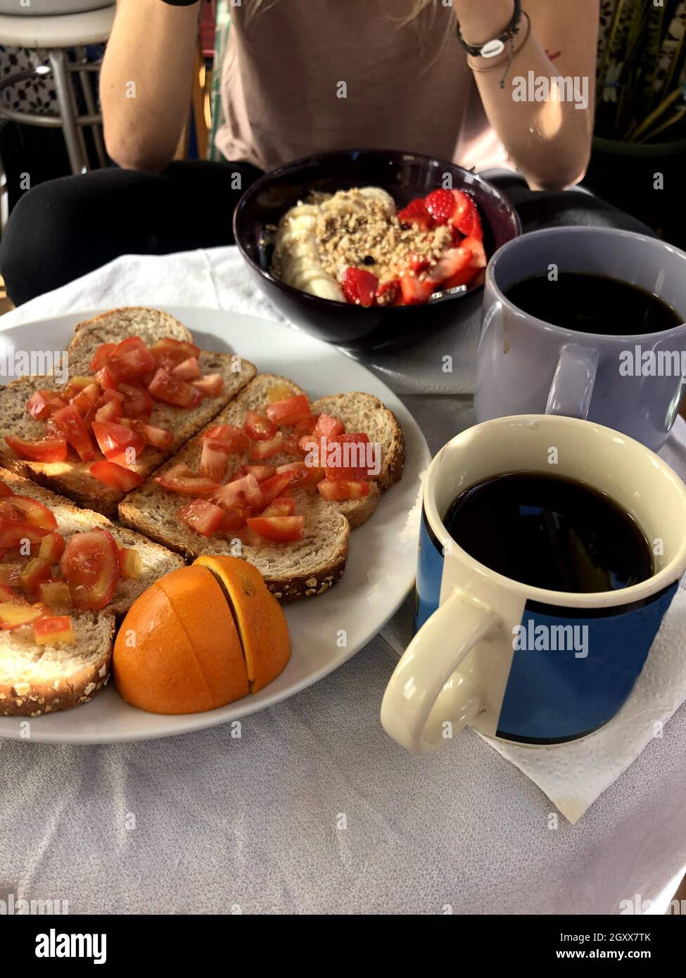 Frau genießt das Frühstück mit Kaffee, Toast mit Tomaten und einer Porridge Schüssel mit Bananen und Erdbeeren Stockfoto