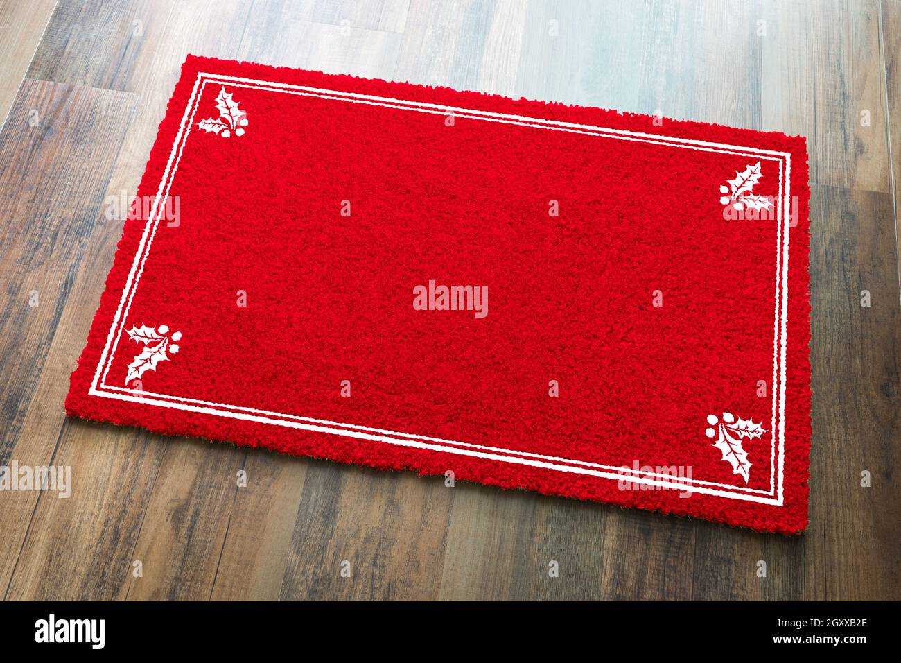 Leere Holiday Red Welcome Matte Mit Holly Ecken Auf Holzboden Hintergrund. Stockfoto