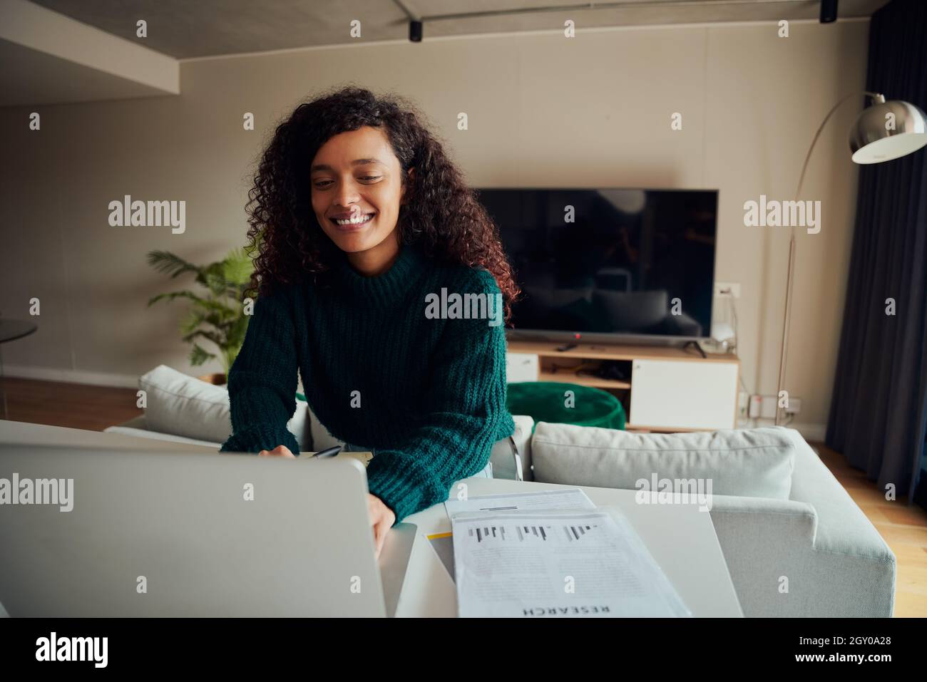 Erwachsene Mischling Frau lächelnd, während sie auf einem Laptop an der Küchentheke sitzt Stockfoto