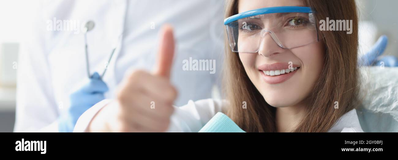 Junge Frau in einer Sicherheitsbrille, die auf einem Zahnarztstuhl sitzt und den Daumen nach oben zeigt Stockfoto