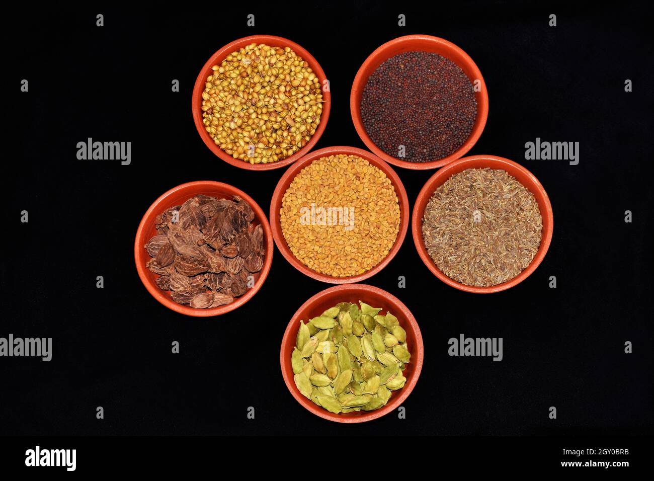 Draufsicht auf indische Gewürze, Fenugreek-Samen, Koriandersamen, Cardamom-Black, Cardamom-Green, Kreuzkümmelkerne, Senfkerne in Tonschüsseln auf schwarzem Hintergrund Stockfoto