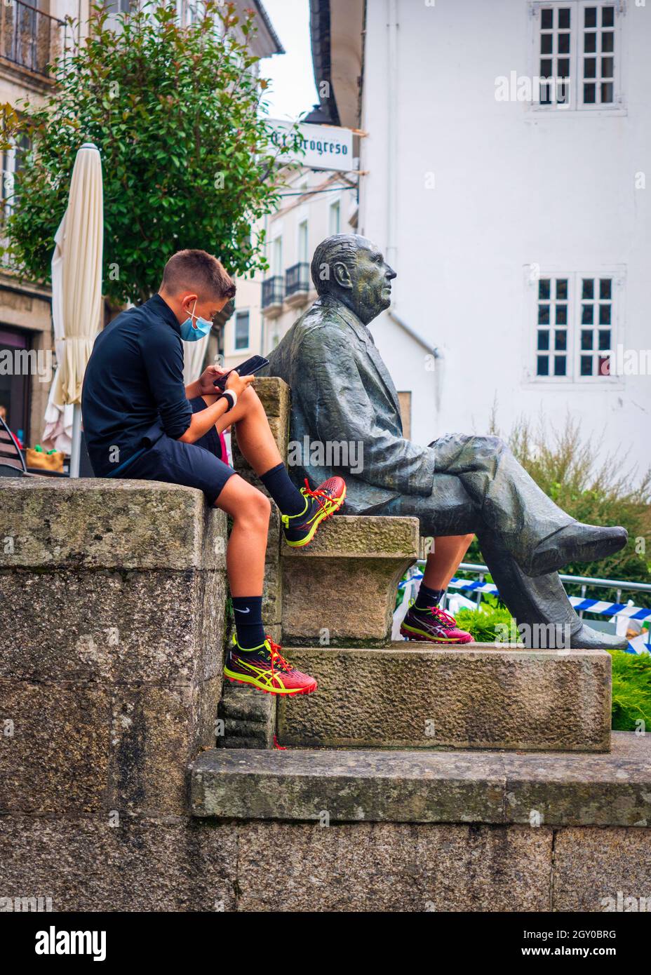 Mondoñedo, Lugo, Spanien. 07,27,2021. Der Junge benutzt sein Handy, das neben der Statue des berühmten Schriftstellers Alvaro Cunqueiro auf dem platz sitzt Stockfoto