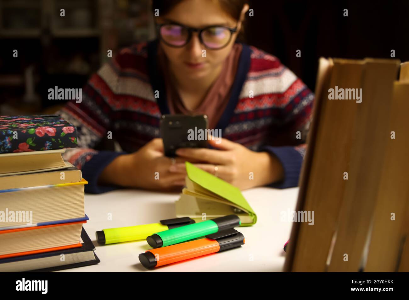 Verschwommener Hintergrund des Bildungs- und Lernkonzepts. Junge lächelnde Frau studiert nachts. Millennial Student Scrolling Phone. Prüfungsvorbereitung. Bücher Stockfoto