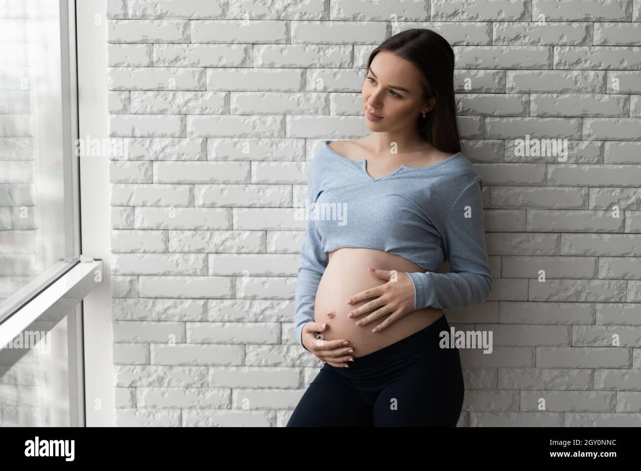 Porträt einer jungen Schwangeren. Steht an einer Ziegelwand und blickt aus dem Fenster. Stockfoto