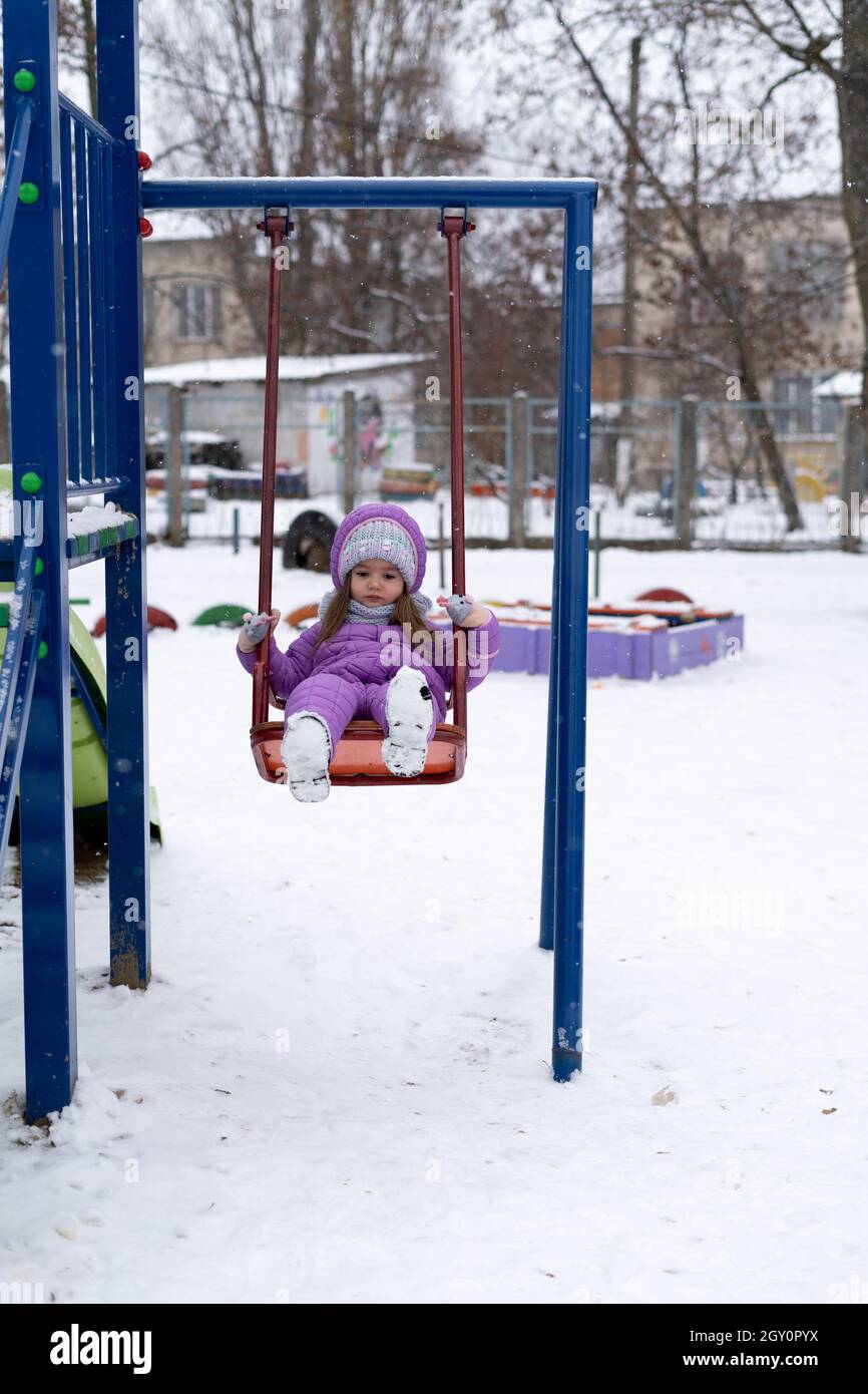 Im Winter, während eines Schneefalls, sitzt ein kleines Mädchen im Vorschulalter hinter einer Schaukel und Schlittschuhe. Stockfoto