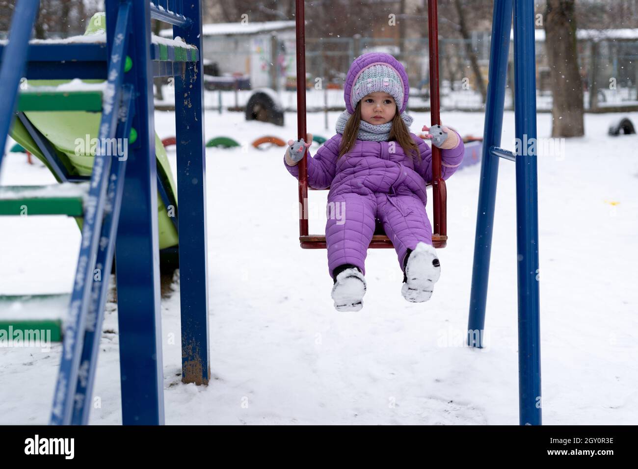 Kinderspielplatz im Winter. Ein kleines Mädchen fährt während eines Schneefalls auf einer Schaukel. Stockfoto