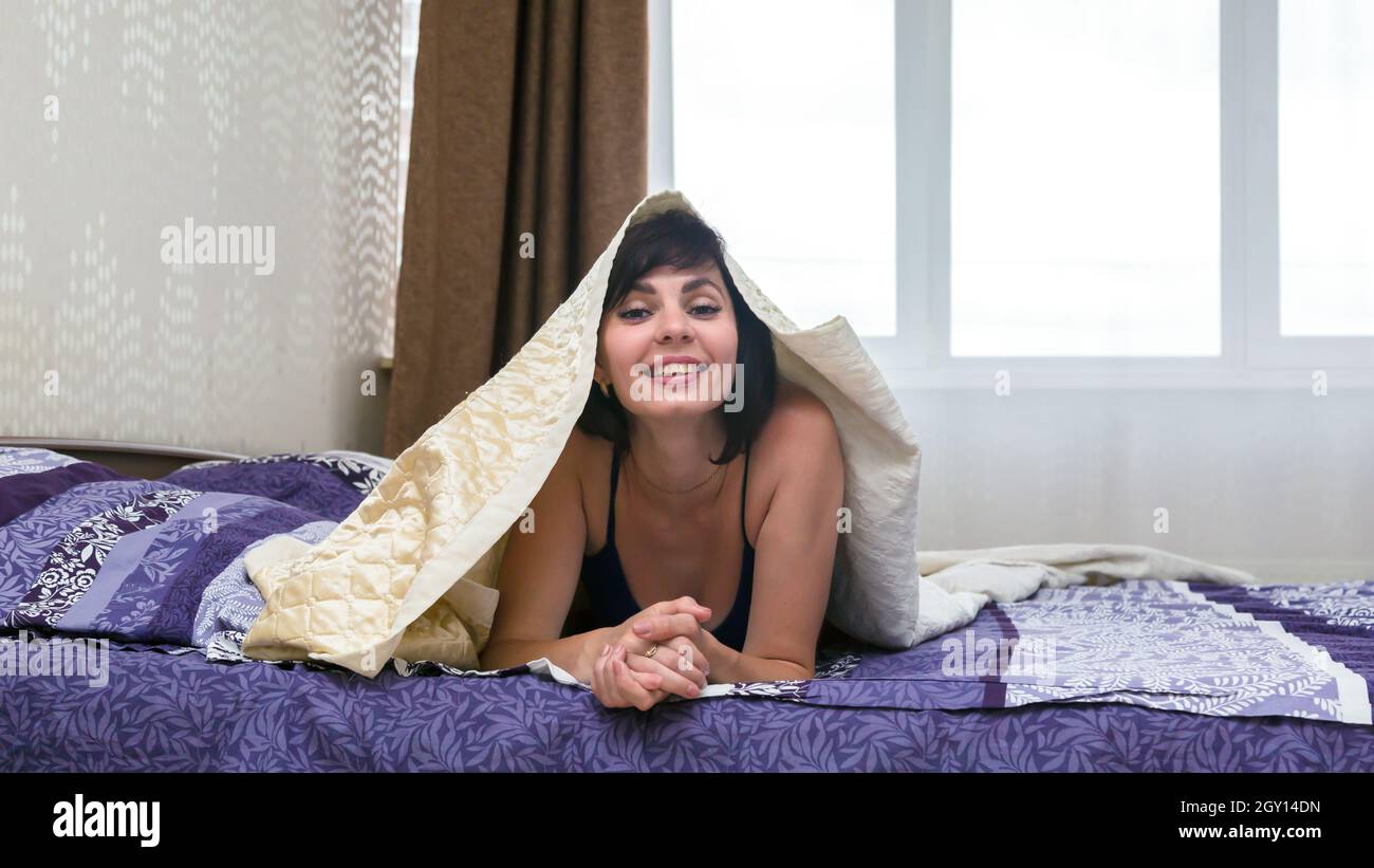Eine fröhliche Brünette liegt auf dem Bett unter weißem Karli. Stockfoto